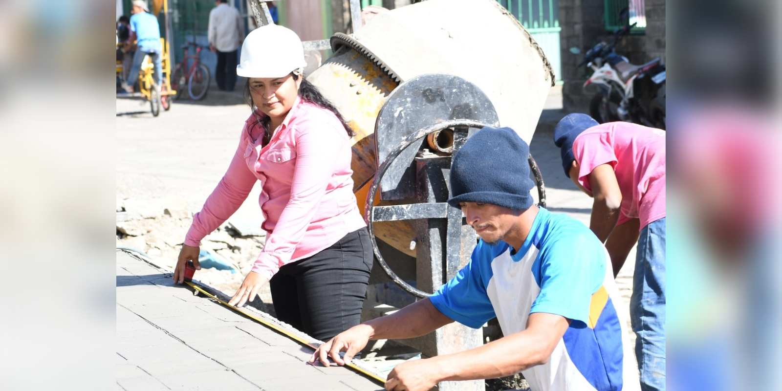 Madeling Benavides supervisa y mide un área de construcción junto a un trabajador, con una mezcladora de cemento al fondo, mostrando su liderazgo en el proyecto.